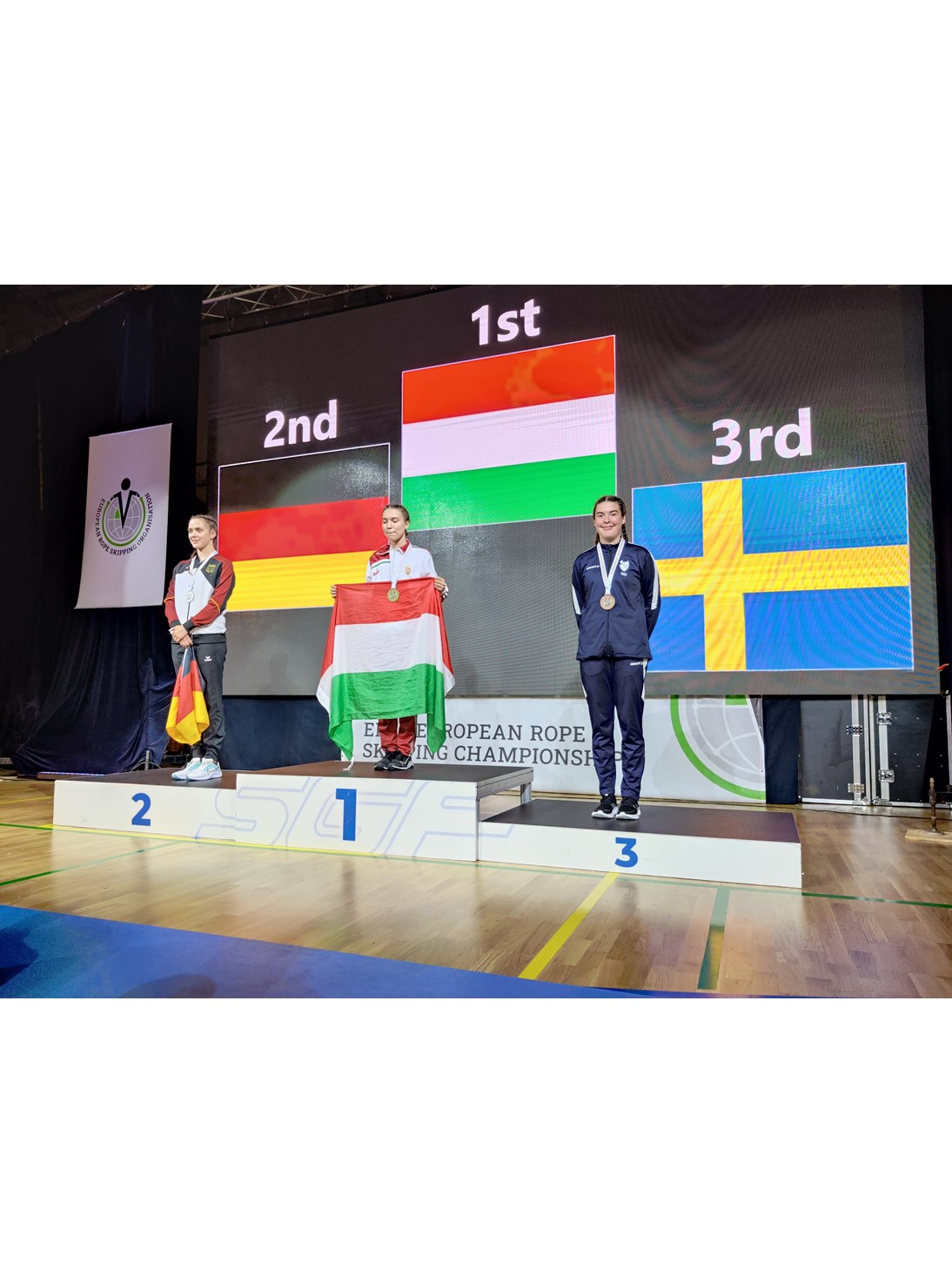 Prispall med svensk tävlande på 3:e plats. Landsflaggor i bakgrunden.